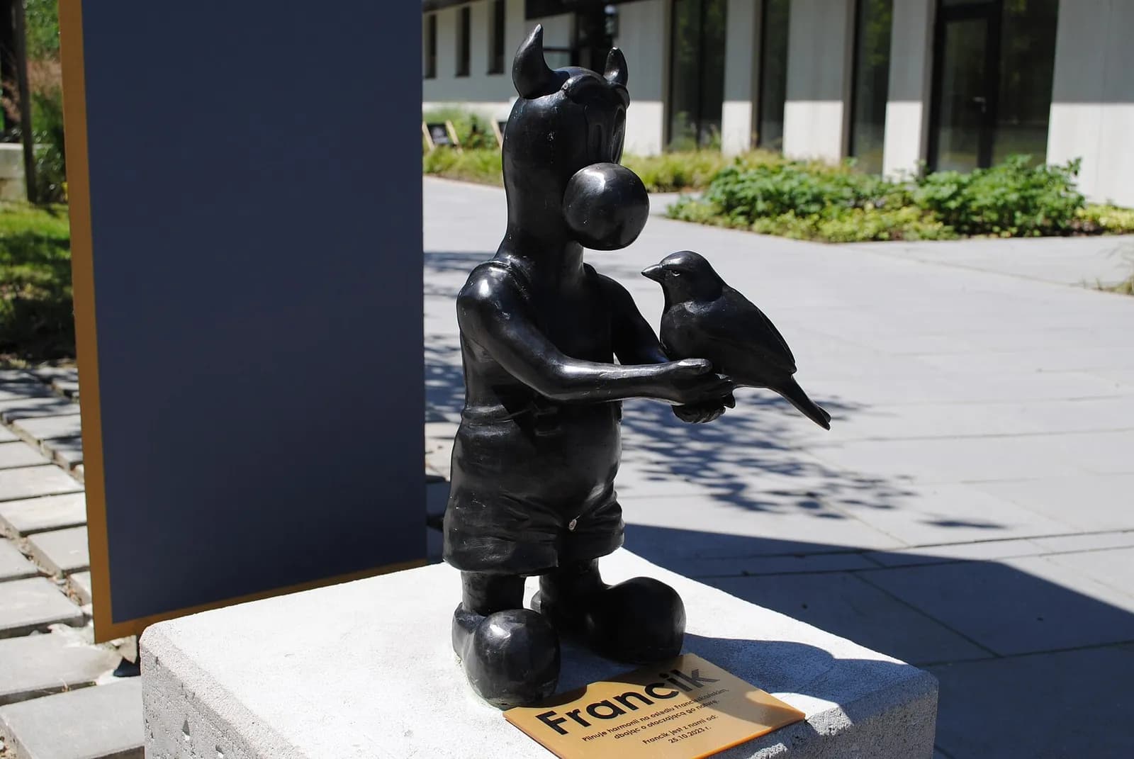 A black statue of a cartoonish figure with horns and a bubble coming from its mouth, holding a small bird in its hands, sitting on a concrete block with a plaque in front of it.