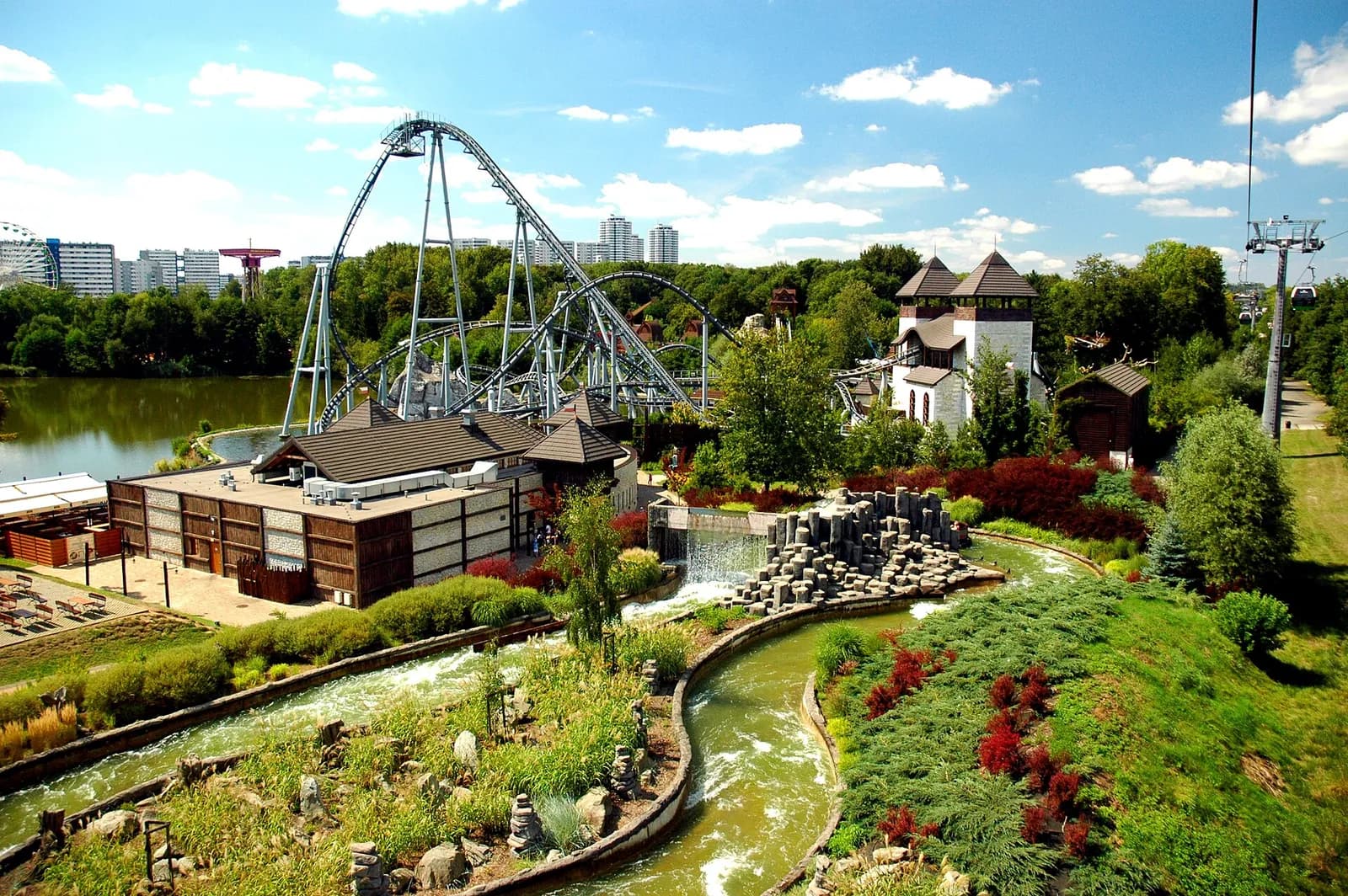 An amusement park features a large roller coaster over a lake, with a winding river ride and a small waterfall in the foreground, and a cable car line on the right.